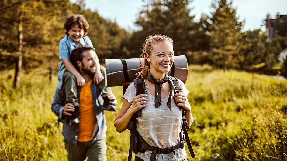 Eine jüngere Frau wandert mit Rucksack, versetzt dahinter läuft ein Mann mit einen kleinen Jungen auf den Schultern